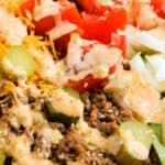 A close-up of a salad with seasoned ground beef, shredded cheese, sliced pickles, tomatoes, onions, and a creamy dressing on a bed of lettuce.