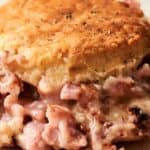 Close-up of a savory biscuit topped pot pie, featuring a golden brown biscuit crust with a creamy filling of diced ham and sauce visible underneath. A knife is placed beside it on a plate.