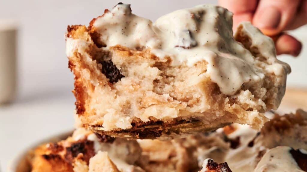 Close-up of someone holding a spoonful of bread pudding topped with creamy sauce.