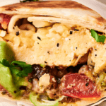 Close-up of a wrapped burrito filled with scrambled eggs, tomatoes, avocado, cheese, and herbs.