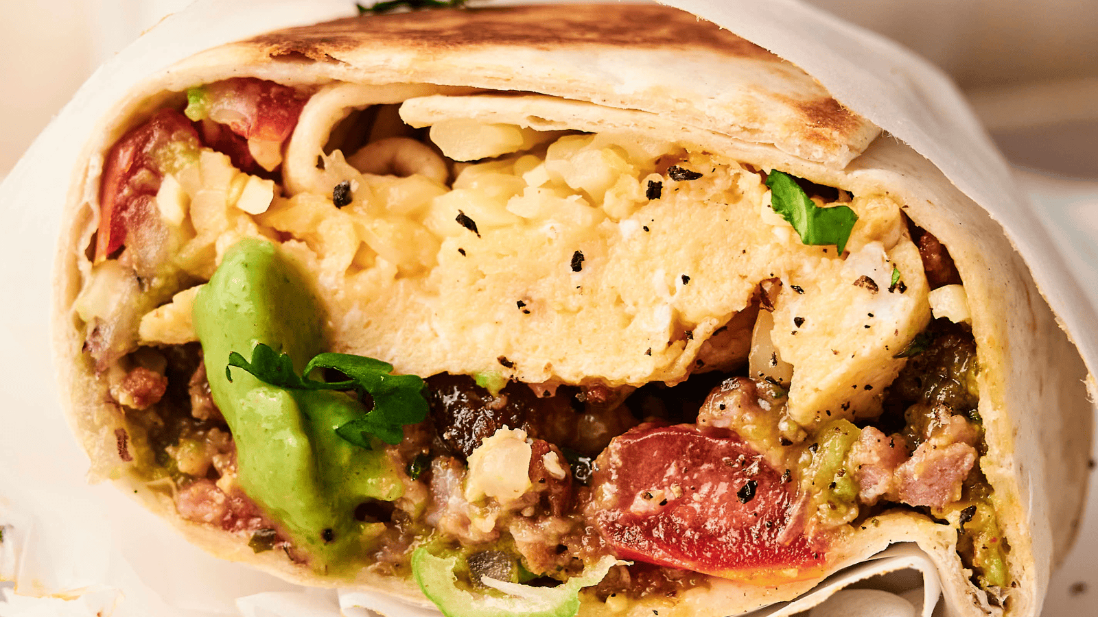 Close-up of a burrito cut in half, showing fillings of scrambled eggs, avocado, tomatoes, cheese, and herbs wrapped in a tortilla.