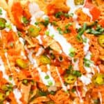 Close-up of nachos topped with melted cheese, sliced jalapeños, chopped green onions, and drizzled with sour cream and hot sauce.