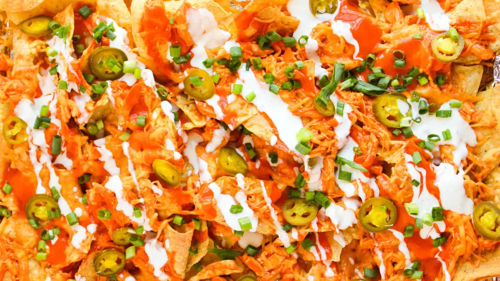 Close-up of loaded Buffalo chicken nachos piled high with shredded chicken, jalapeños, cheese, green onions, hot sauce, and a dollop of sour cream.