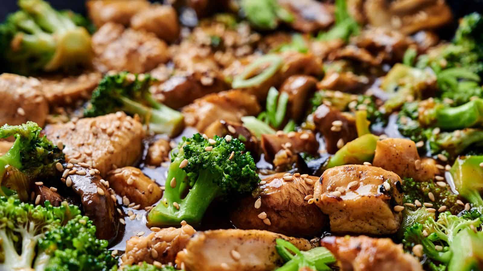 Close-up of a stir-fry dish with chicken, broccoli, mushrooms, and sesame seeds, coated in a soy-based sauce.