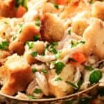 Close-up of a spoon scooping savory stuffing with chunks of bread, peas, carrots, and herbs.