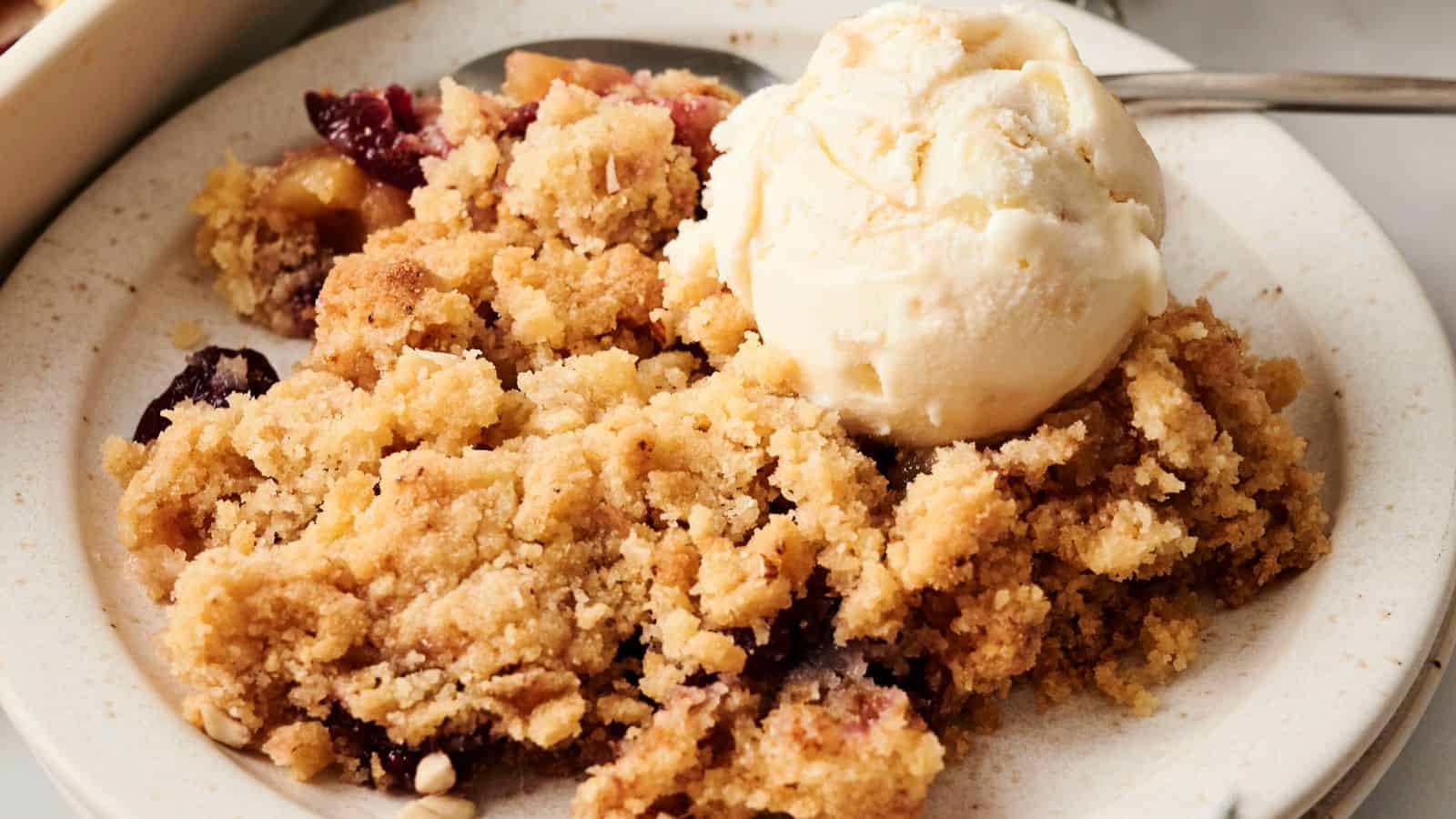 A plate of fruit crumble topped with a scoop of vanilla ice cream, accompanied by a spoon.