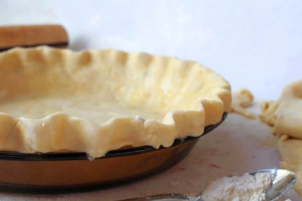 Unbaked pie crust in a brown pie dish on a countertop, with a rolling pin and flour-dusted spoon nearby.