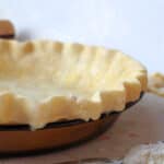 Unbaked pie crust in a brown pie dish on a countertop, with a rolling pin and flour-dusted spoon nearby.