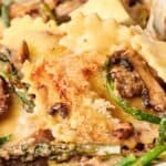 Close-up of a pasta dish featuring ravioli, mushrooms, asparagus, and melted cheese in a baking dish.