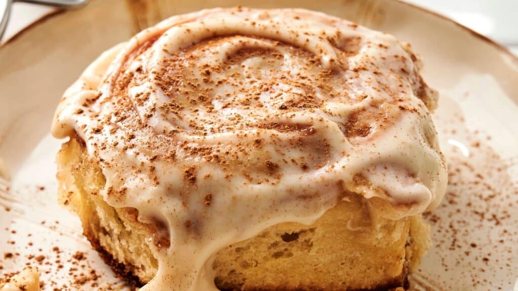 Close-up of a cinnamon roll topped with creamy icing and a sprinkle of cinnamon on a beige plate.