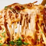 Close-up of cheesy baked spaghetti being lifted with a fork, featuring melted cheese, tomato sauce, and garnished with parsley.