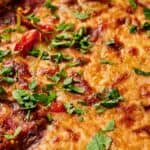 Close-up of a baked dish topped with melted cheese and garnished with chopped herbs, featuring visible pieces of tomato and green bell pepper.