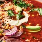 A bowl of tomato-based soup topped with avocado slices, cilantro, red onion, corn, shredded cheese, and a dollop of sour cream.