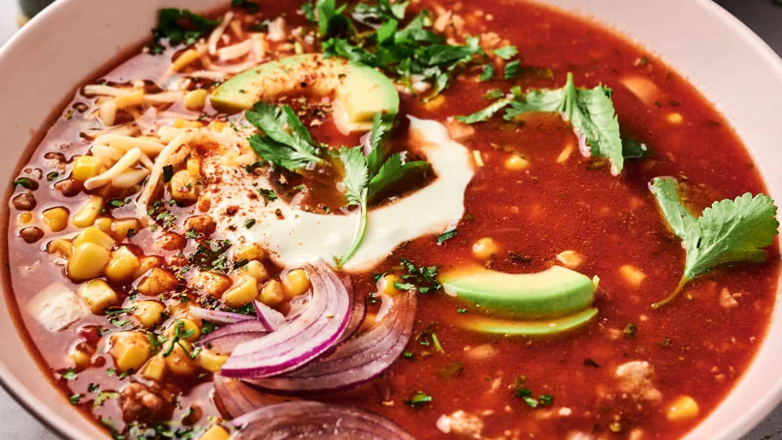 A bowl of red soup topped with avocado slices, sour cream, cilantro, sliced onions, corn, and shredded cheese.