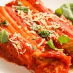 Plate of baked cannelloni filled with cheese, topped with tomato sauce, grated cheese, and fresh basil leaves.