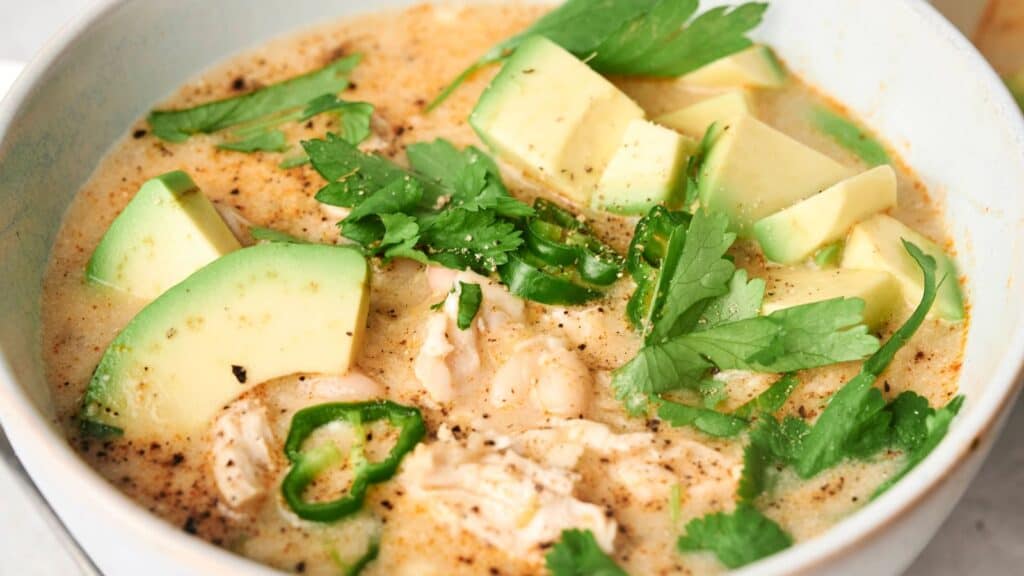 A bowl of creamy chicken soup topped with avocado slices, jalapeño slices, and fresh cilantro.