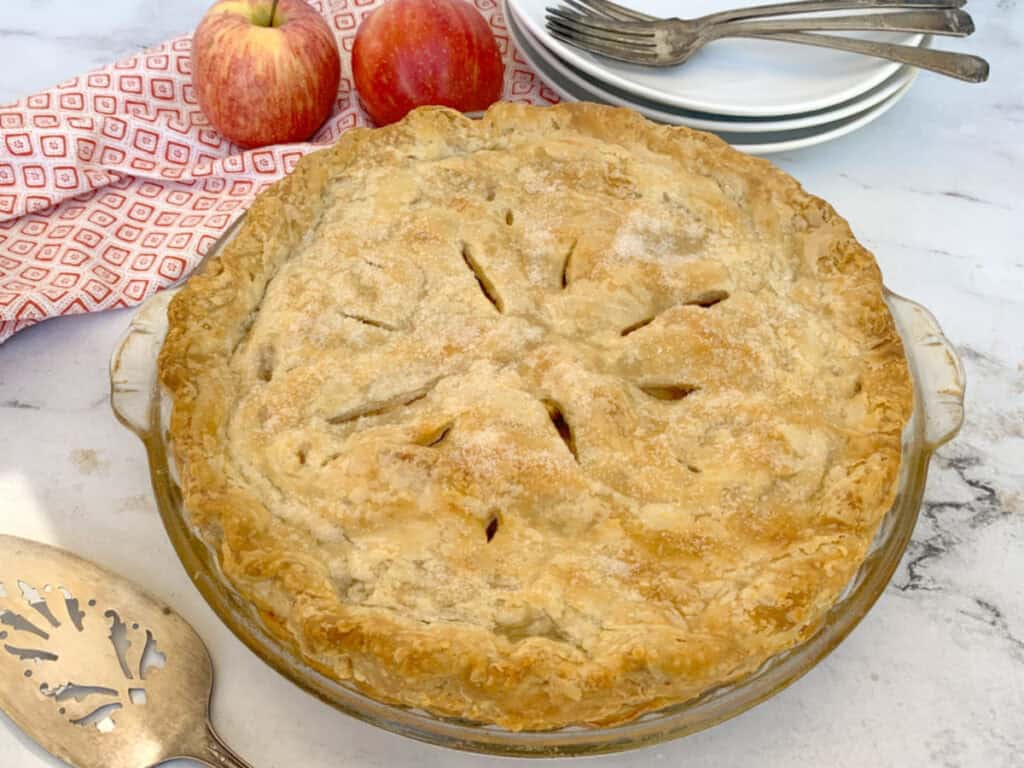 A freshly baked apple pie in a glass dish on a marble countertop, accompanied by two apples, a red patterned cloth, and stacked white plates with forks beside it.