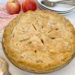 A freshly baked apple pie in a glass dish on a marble countertop, accompanied by two apples, a red patterned cloth, and stacked white plates with forks beside it.