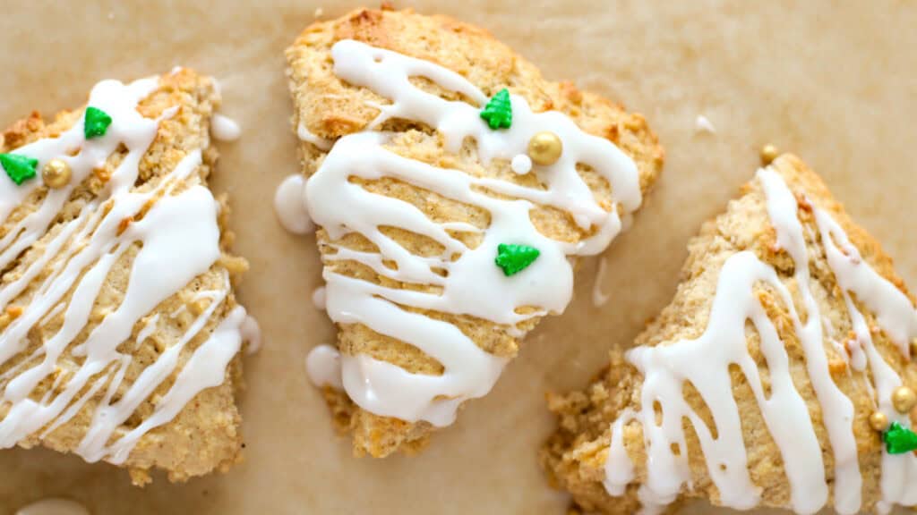 Three triangular scones with white icing drizzles and small green decorations on a parchment-lined surface.