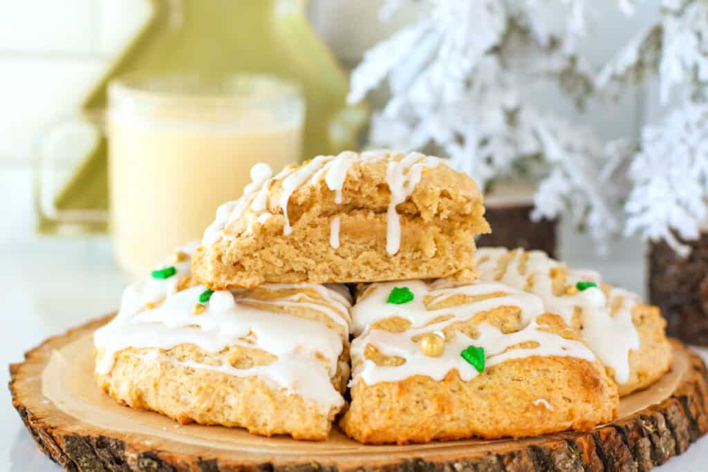 Three frosted scones with festive sprinkles are stacked on a wooden platter. A glass of eggnog and a snowy decoration are blurred in the background.