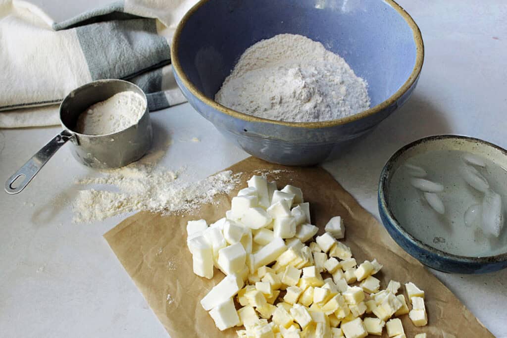 Bowls of flour and ice water, cubes of butter on parchment, measuring cup with flour, and a towel on a white surface.