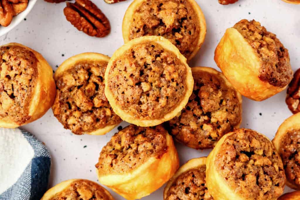 A close-up of several pecan tarts arranged on a white surface, with some whole pecans visible in the background.