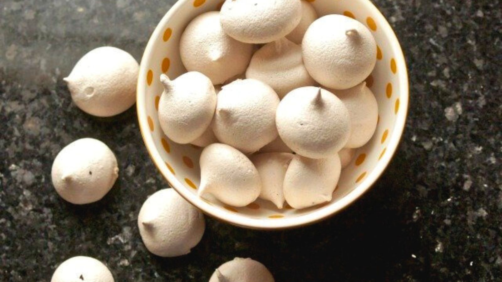 A bowl filled with mocha meringue cookies, some scattered on a dark textured surface.