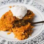 A slice of carrot cake on a patterned plate, topped with a dollop of whipped cream, with a fork resting beside it.