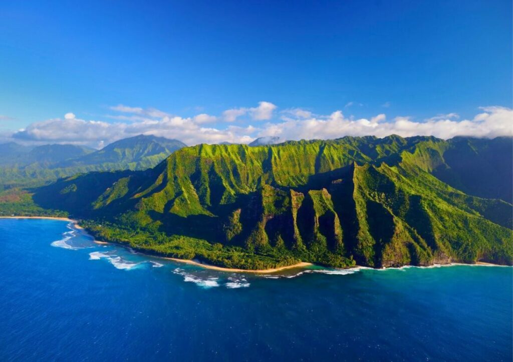 Aerial view of lush green cliffs by the ocean under a clear blue sky, with waves gently hitting the shore—a stunning sight that captures one of the top things to do in Kauai.