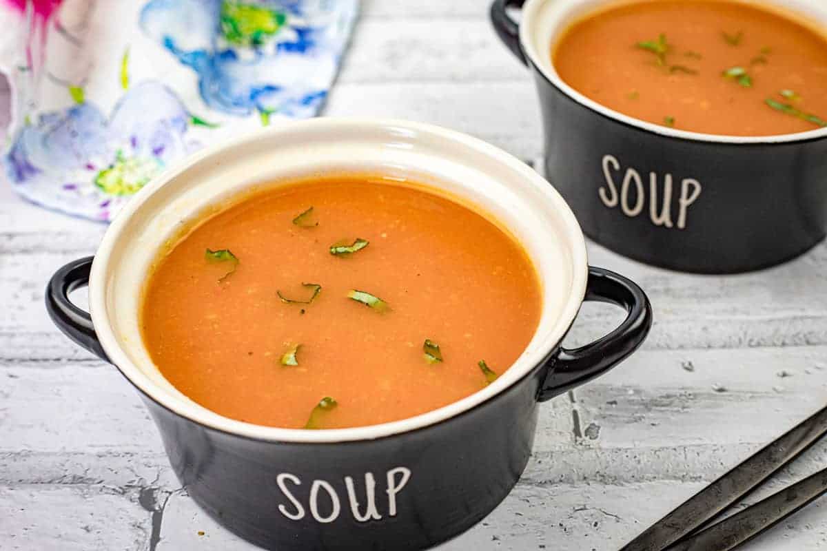 Two bowls of old fashioned tomato soup with chopped herbs on top, placed on a white wooden surface.