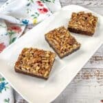 Three pecan-topped bars on a white rectangular plate, placed on a white wooden surface with a floral-patterned cloth nearby.