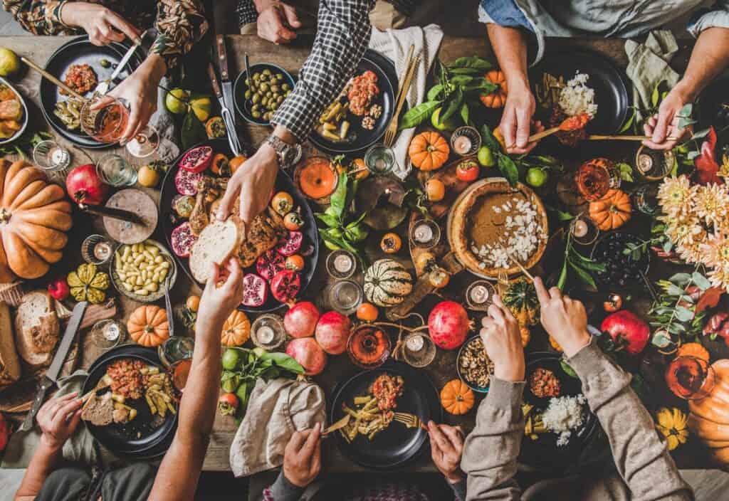 A table brimming with a variety of food, including pumpkins, pomegranates, and dishes being shared by multiple hands, paints the perfect picture of intimate small gatherings and festive cheer.