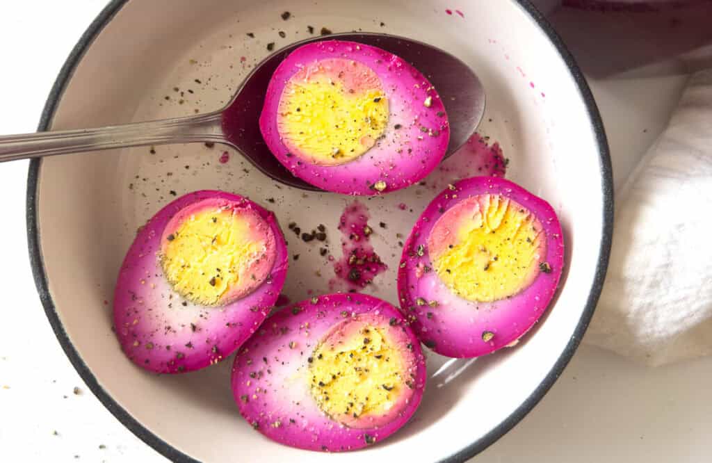 Sliced pickled eggs with bright pink edges, topped with pepper, in a bowl with a spoon.