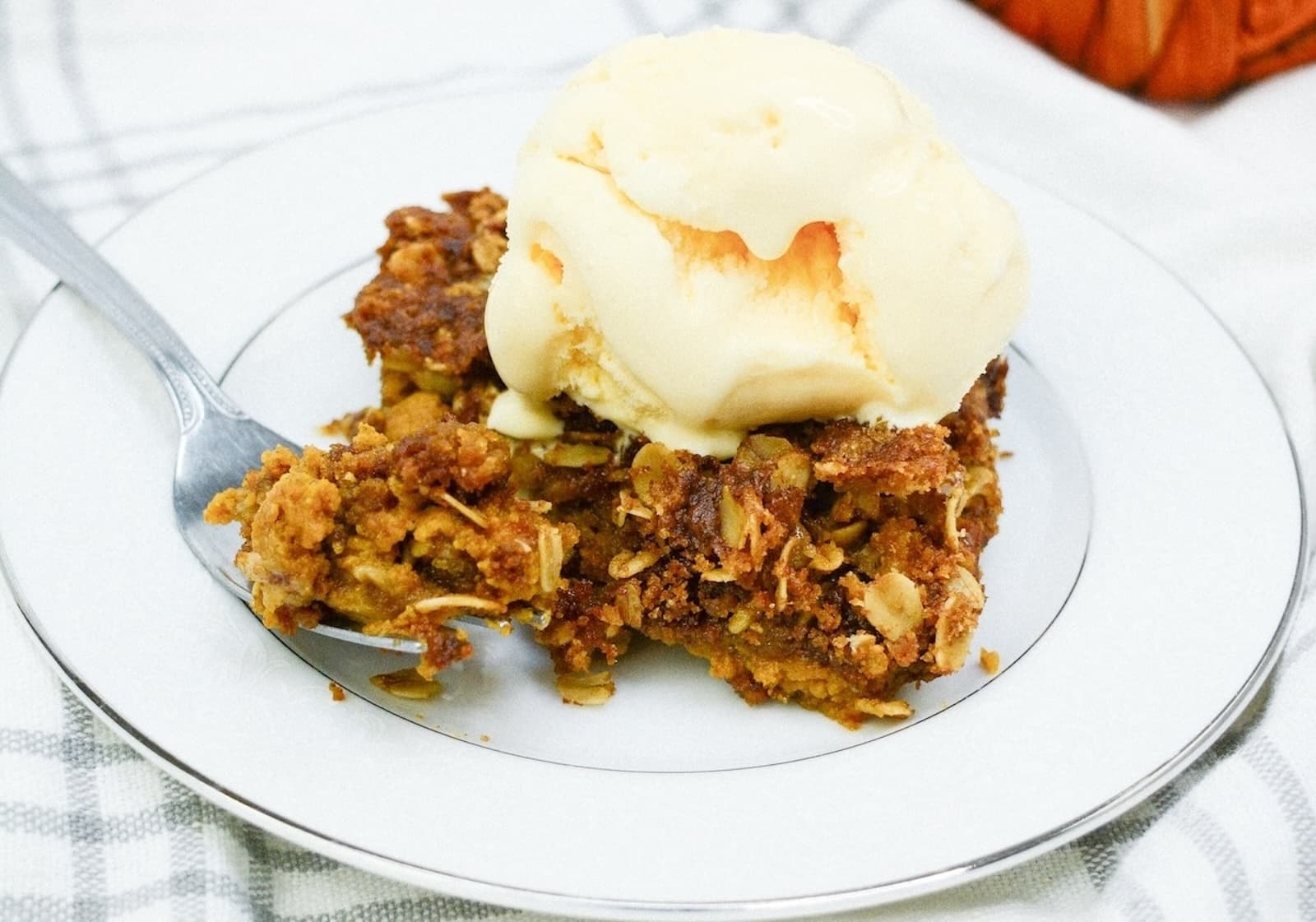 A piece of pumpkin crisp topped with a scoop of vanilla ice cream on a white plate, placed on a striped cloth.