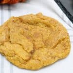 A single pumpkin cookie on a checkered cloth, with another cookie partially visible on a black tray in the background.