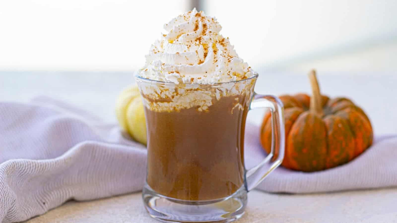 A glass mug of pumpkin spice latte topped with whipped cream and cinnamon, set on a table with a white cloth. Two small pumpkins are in the background.