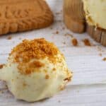 White chocolate pumpkin truffle topped with cookie crumbs, next to whole cookies on a white wooden surface.