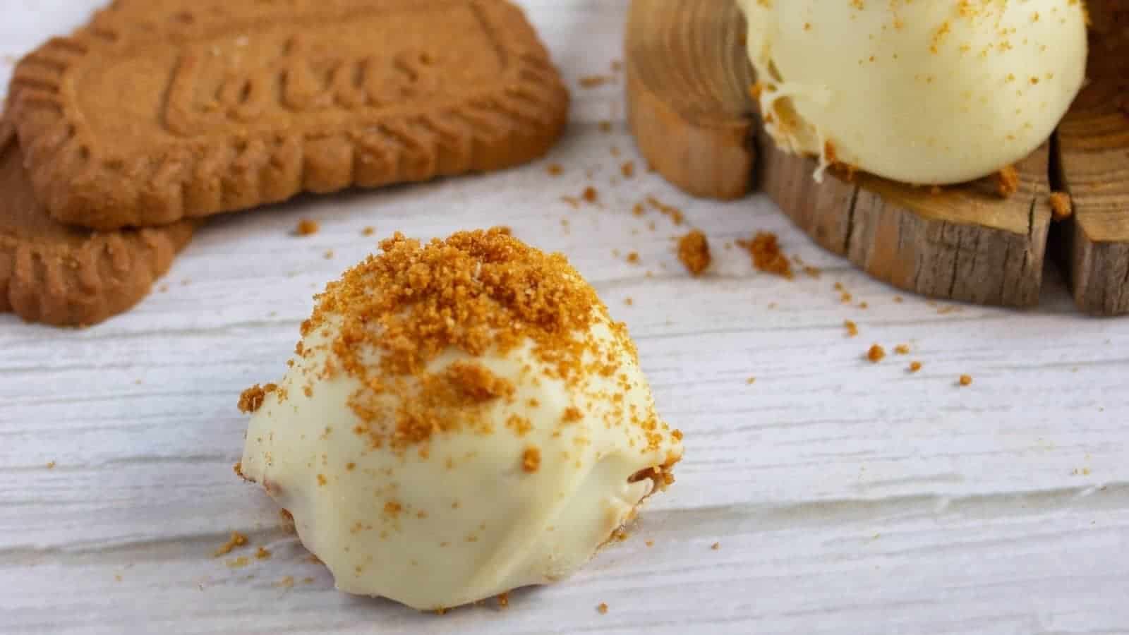 White chocolate pumpkin truffle topped with cookie crumbs, next to whole cookies on a white wooden surface.