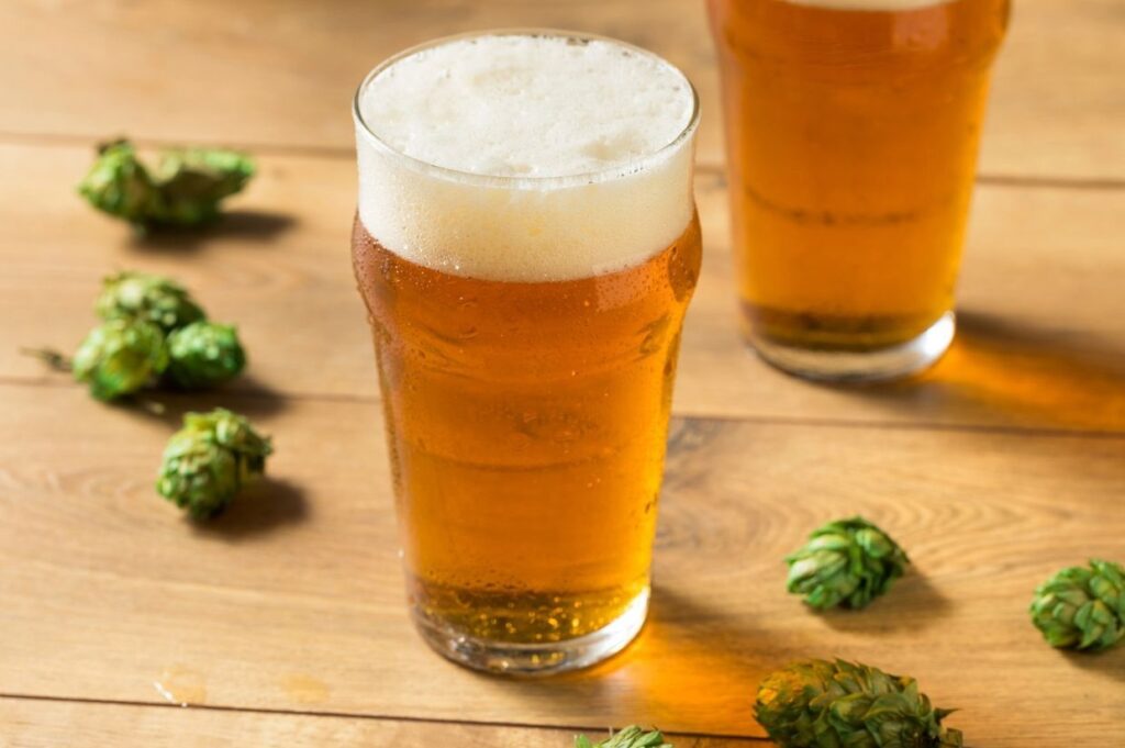 Two glasses of amber beer with frothy heads on a wooden table, surrounded by scattered green hops.