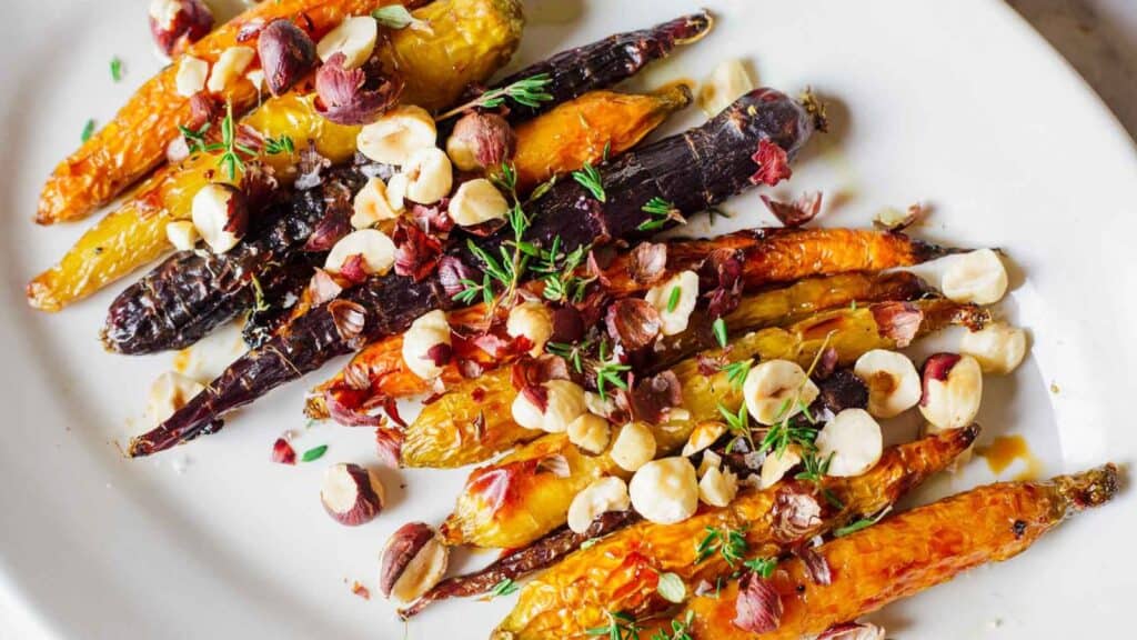 Roasted colorful carrots topped with hazelnuts and herbs on a white plate.