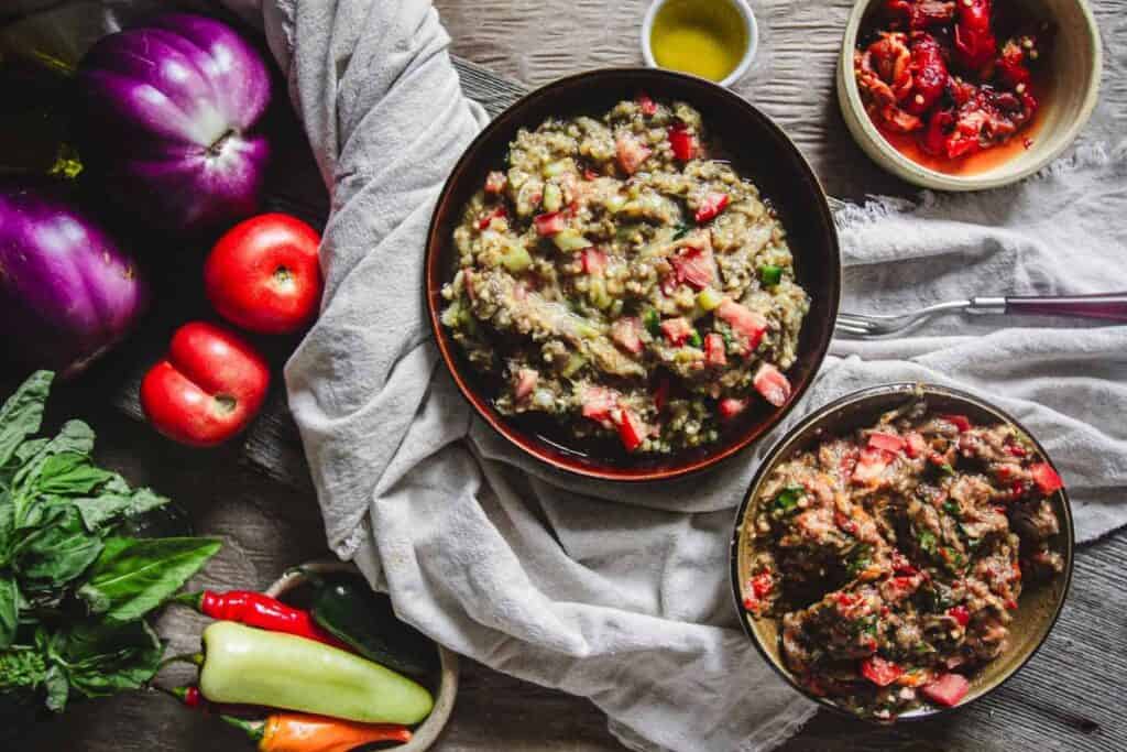 Two bowls of vegetable stew on a cloth with a side of peppers and basil. Apples and eggplants are visible on the left. A small dish of oil and a bowl of tomatoes are on the right.