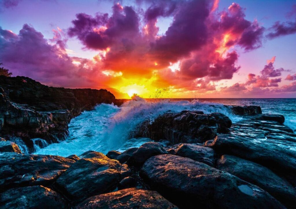 Waves crash against Kauai's rocky cliffs at sunset, with vibrant orange and purple clouds filling the sky—a mesmerizing sight and one of the top things to do in Kauai.