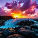 Waves crash against Kauai's rocky cliffs at sunset, with vibrant orange and purple clouds filling the sky—a mesmerizing sight and one of the top things to do in Kauai.