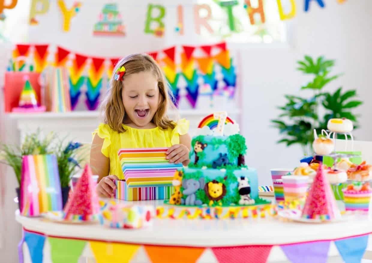 A young girl in a yellow dress admires a gift at a colorful birthday party table with a rainbow-themed cake, decorations, and party hats.