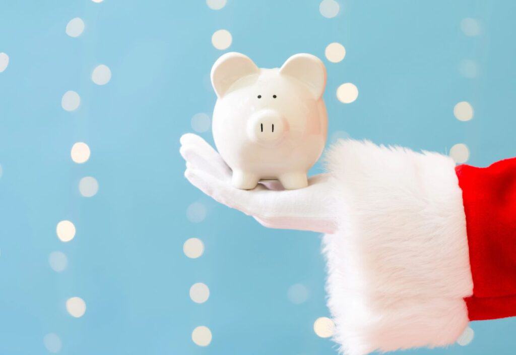 A Santa Claus hand holding a white piggy bank against a blue background with bokeh lights.