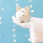 A Santa Claus hand holding a white piggy bank against a blue background with bokeh lights.