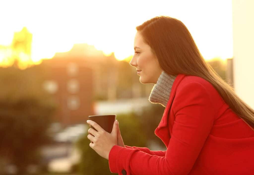 A woman in a red coat holds a mug, gazing into the distance during a sunset, finding solace to help get through the holidays.