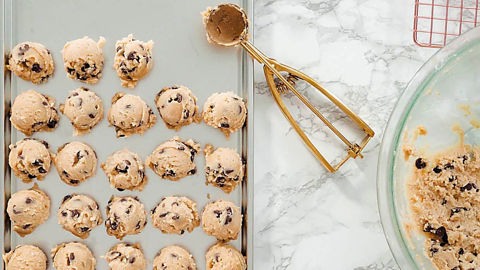 A baking tray with evenly spaced scoops of chocolate chip cookie dough, a gold ice cream scoop, and a mixing bowl with leftover dough on a marble surface—perfectly prepped for chilling cookie dough before baking.