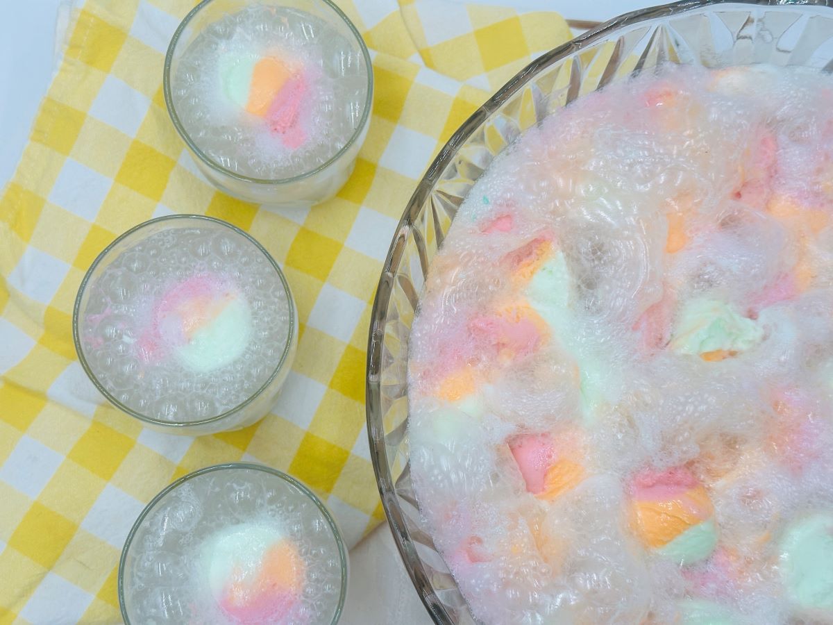 Bowl and glasses of rainbow sherbet punch with vibrant foam on a yellow plaid tablecloth.