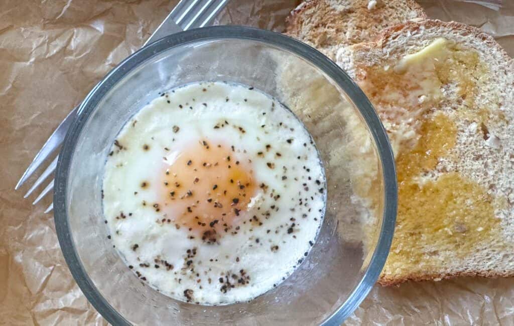 A glass bowl with a baked egg seasoned with pepper next to a slice of buttered toast and a fork on parchment paper.